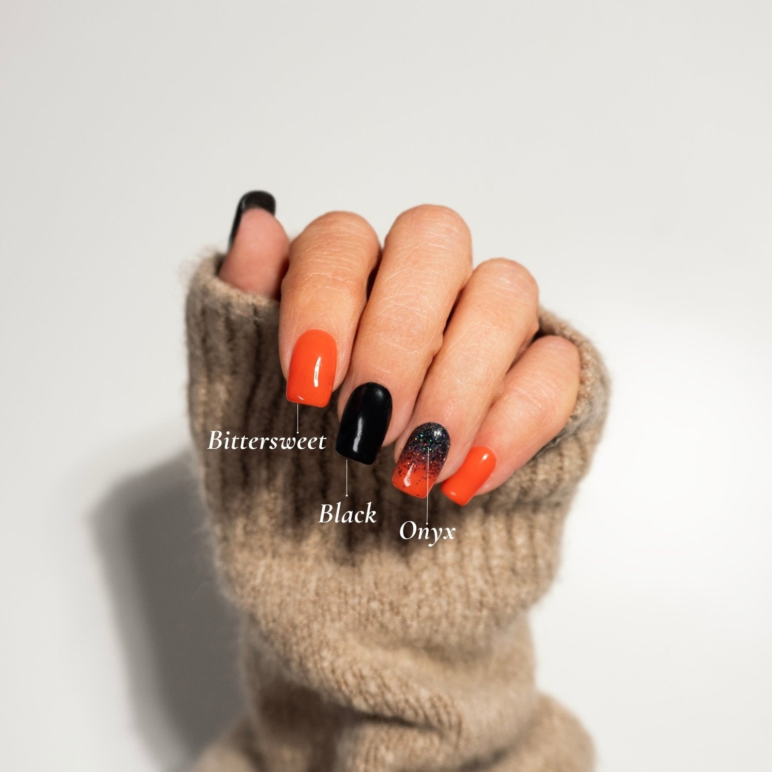 Hand mit auffälligem Nail-Design in den Farben 'Bittersweet' (knalliges Orange), 'Black' (tiefes Schwarz) und 'Onyx' (schwarz mit Glitzer) von DIPDRIP. Das kreative Design kombiniert kräftige Farben und funkelnde Akzente, perfekt hervorgehoben durch den neutralen Hintergrund und einen beigen Pullover