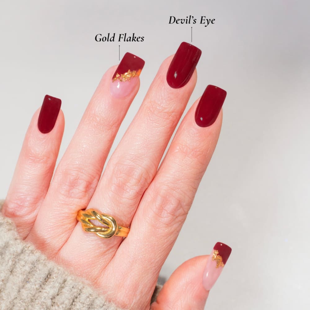 Hand mit langen, eleganten Nägeln, lackiert in einem tiefen Rotton namens 'Devil's Eye', kombiniert mit transparenten Nägeln und goldenen Akzenten ('Gold Flakes'). Die Nägel sind in einem stilvollen Dipping-Powder-Design gestaltet, ergänzt durch einen goldenen Ring und einen beigen Strickpullover im Hintergrund.