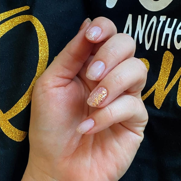Zartrosa Nägel mit Goldglitzer an den Spitzen vor schwarzem Shirt mit Goldschrift