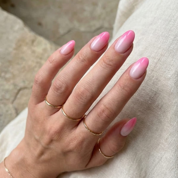 Mandelnägel im rosa Farbverlauf, Hand mit dünnen Goldringen auf hellem Stoff